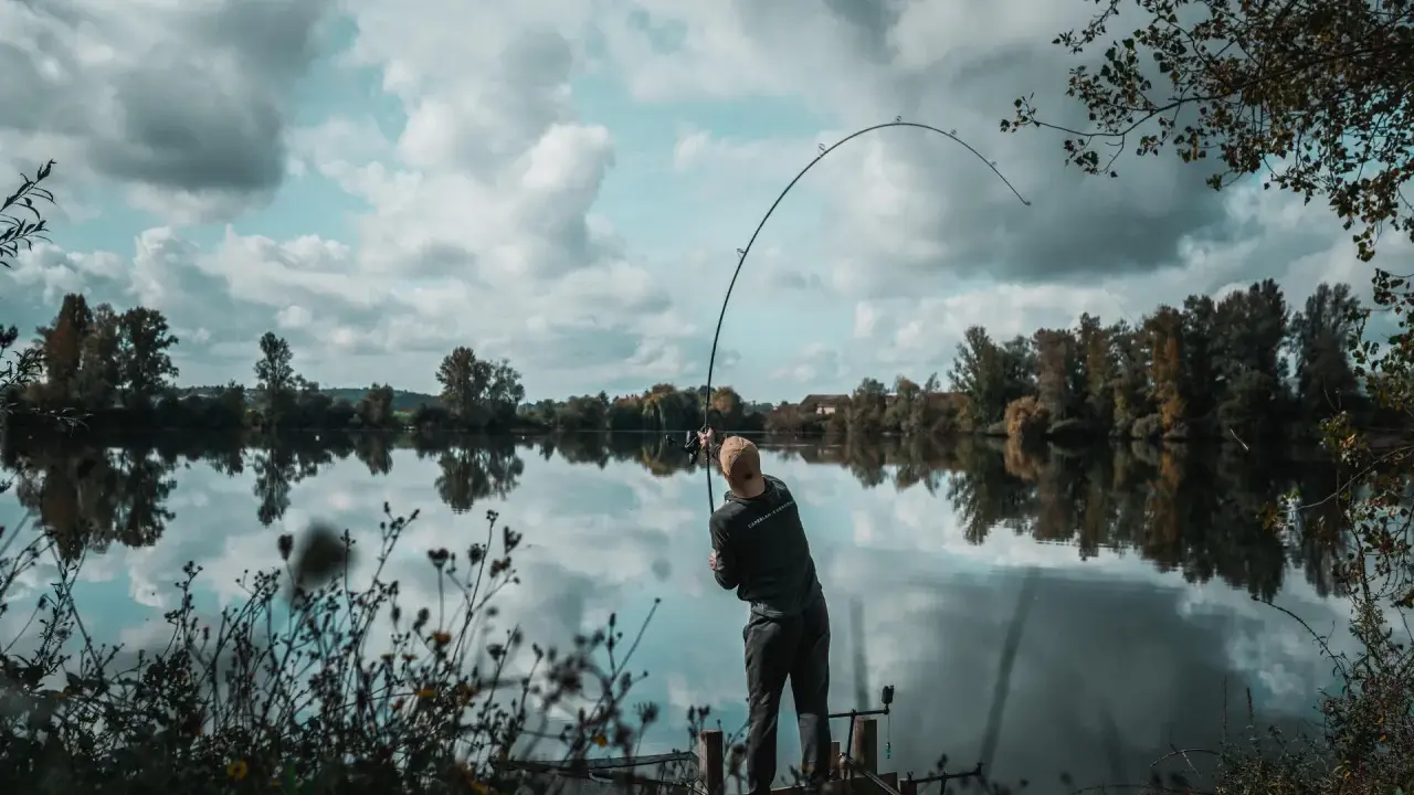 Wędkarz z napiętą wędką, łowiący ryby zgodnie z księżycowym kalendarzem brań. Spokojna tafla wody odbija chmury.