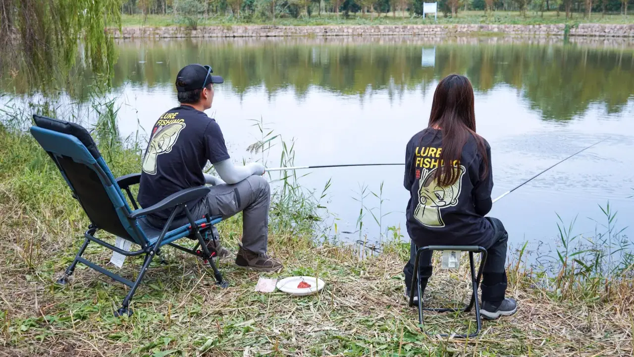 Para wędkująca nad jeziorem, w koszulkach z napisem "Lure Fishing". Cieszą się spokojem, choć ich hobby to czysta komercja.