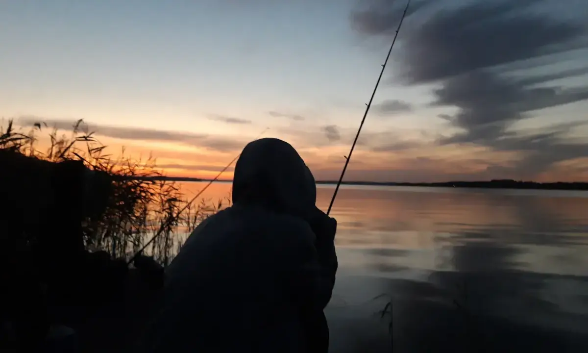Zachód słońca nad jeziorem, wędkarz z wędką czeka na branie. Idealna karta rybacka.