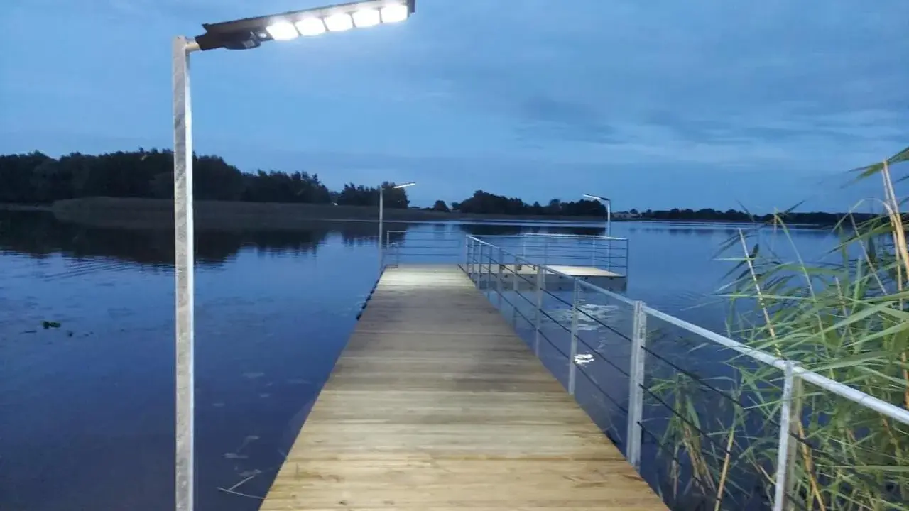 Drewniany pomost nad jeziorem, oświetlony lampami, zachęca do spaceru. Czy pomost nad jeziorem może być prywatny? Ten wygląda na dostępny dla wszystkich.