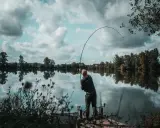 Wędkarz z napiętą wędką, łowiący ryby zgodnie z księżycowym kalendarzem brań. Spokojna tafla wody odbija chmury.