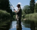 Wędkarz w wodzie, z wędką w ręku, czeka na branie w spokojnym jeziorze rokola. Otaczają go bujna zieleń i błękitne niebo.