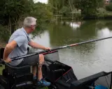 Mężczyzna siedzi nad wodą, przygotowując wędkę na grunt. Wędka jest długa, z widocznym napisem "CARPCOVER".