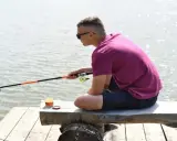 Młody mężczyzna w okularach przeciwsłonecznych siedzi na drewnianej ławce nad wodą, trzymając wędkę. Czy wędkarstwo to sport? On chyba tak uważa.