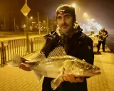 Szczecin rzeka nocą. Wędkarz z okazałym sandaczem na tle oświetlonej promenady.