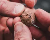 Dłonie trzymają okrągłą kulkę przynęty wędkarskiej z widocznymi fragmentami.