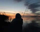 Zachód słońca nad jeziorem, wędkarz z wędką czeka na branie. Idealna karta rybacka.