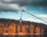 Przynęta z haczykiem wisi na żyłce, gotowa do połowu na jeziorze Skarlińskim. W tle jesienne drzewa i błękitne niebo.