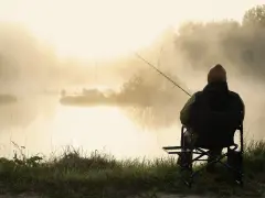 Wędkarz siedzi nad mglistym jeziorem o poranku. Słońce przebija się przez mgłę, tworząc złocistą poświatę.
