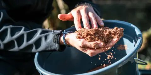 Ręce trzymają zanętę Traper Feeder nad wiaderkiem.