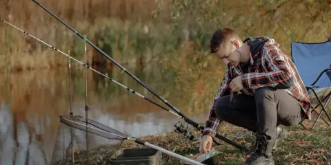 Mężczyzna przygotowuje się do łowienia, sprawdzając przynętę. W tle wędki i siatka, jak zrobić wędkę na grunt to jego cel.