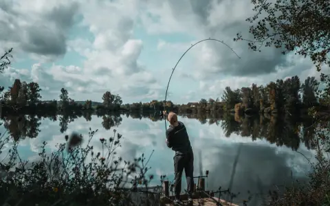 Wędkarz z napiętą wędką, łowiący ryby zgodnie z księżycowym kalendarzem brań. Spokojna tafla wody odbija chmury.
