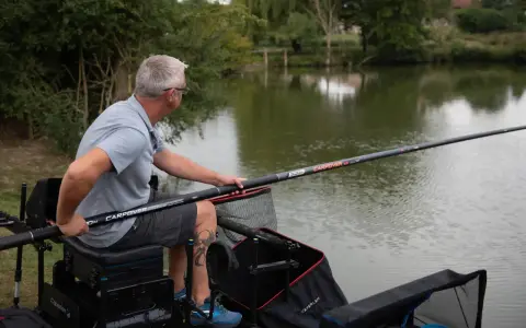 Mężczyzna siedzi nad wodą, przygotowując wędkę na grunt. Wędka jest długa, z widocznym napisem "CARPCOVER".