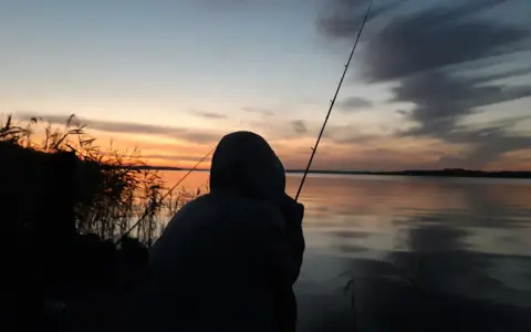 Zachód słońca nad jeziorem, wędkarz z wędką czeka na branie. Idealna karta rybacka.