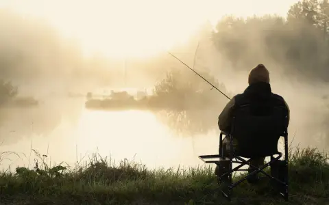 Wędkarz siedzi nad mglistym jeziorem o poranku. Słońce przebija się przez mgłę, tworząc złocistą poświatę.