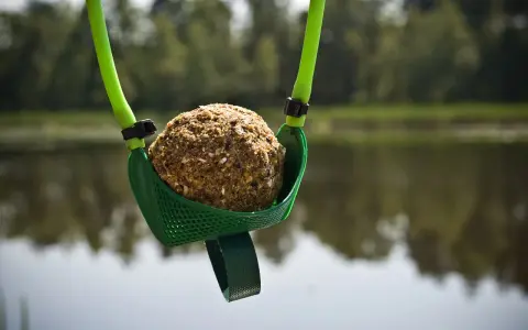 Zielona proca do zanęty z kulką zanętową gotowa do rzutu nad spokojną wodą.