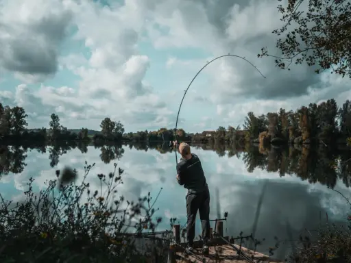 Wędkarz z napiętą wędką, łowiący ryby zgodnie z księżycowym kalendarzem brań. Spokojna tafla wody odbija chmury.