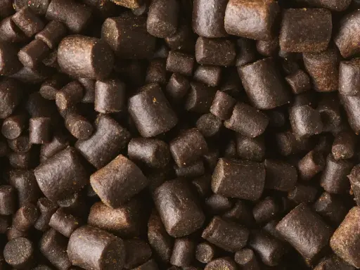 Zbliżenie na ciemnobrązowe granulki zanęty wędkarskiej o nieregularnych kształtach.