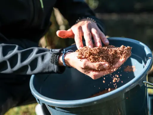 Ręce trzymają zanętę Traper Feeder nad wiaderkiem.