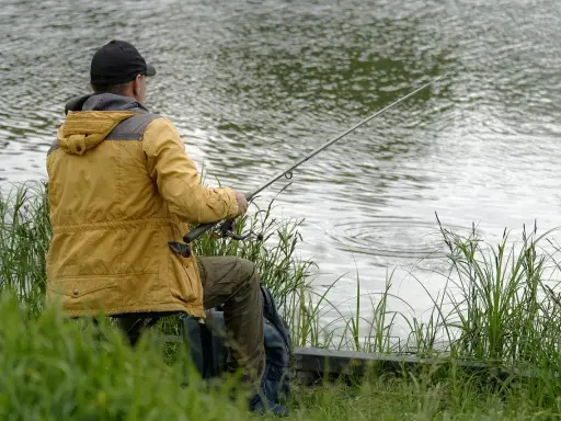 Wędkarz nad jeziorem, przygotowuje zanętę na ryby. Woda faluje, a trawa otacza brzeg.
