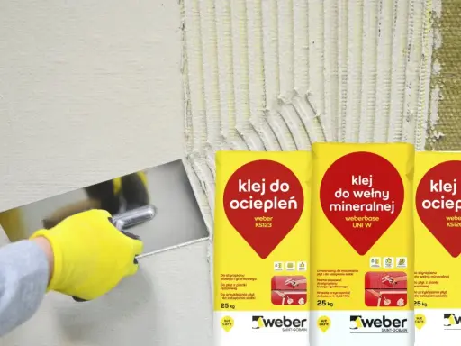 Ręka w żółtej rękawicy nakłada klej do ociepleń na ścianę. Obok worki z klejem do wełny mineralnej i do ociepleń.
