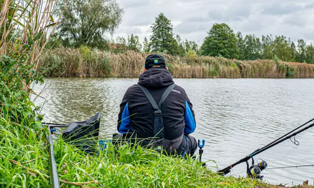 Wędkarz siedzi nad rzeką, przygotowując zanętę na rzekę feeder. W tle trzciny i drzewa.