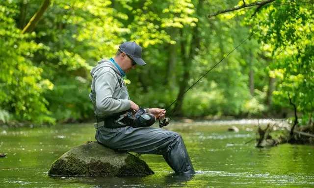 Wędkarz siedzi na kamieniu w zielonej wodzie, przygotowując sprzęt. Spokój nad zalewem Jaraczewo.