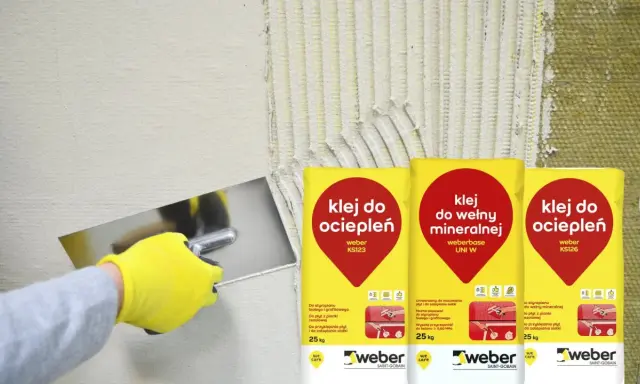 Ręka w żółtej rękawicy nakłada klej do ociepleń na ścianę. Obok worki z klejem do wełny mineralnej i do ociepleń.