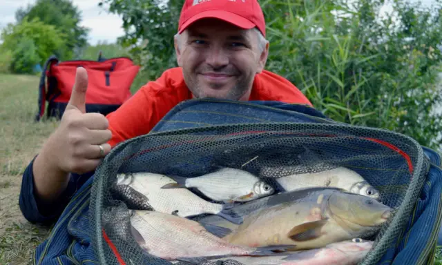 Szczęśliwy wędkarz z okazałym połowem ryb w podbieraku. W tle czerwony plecak i zielona roślinność. Gotowa zanęta do metody zadziałała!