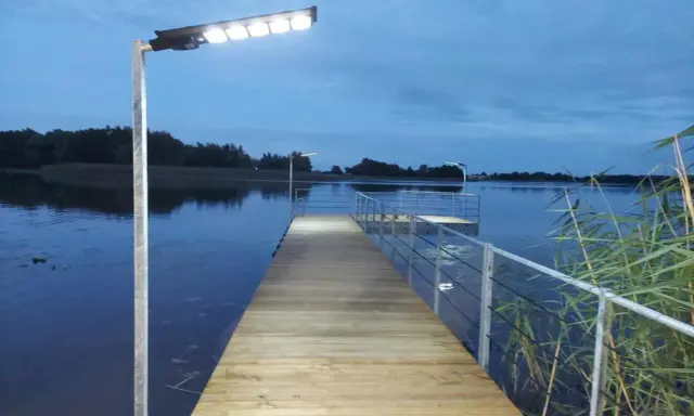 Drewniany pomost nad jeziorem, oświetlony lampami, zachęca do spaceru. Czy pomost nad jeziorem może być prywatny? Ten wygląda na dostępny dla wszystkich.