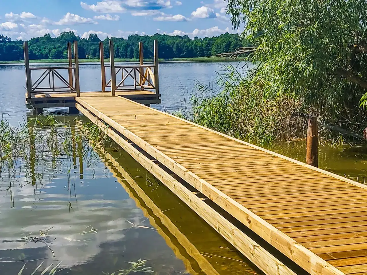 Drewniany pomost na jeziorze zaprasza do spaceru. Woda odbija błękitne niebo i zielone drzewa.