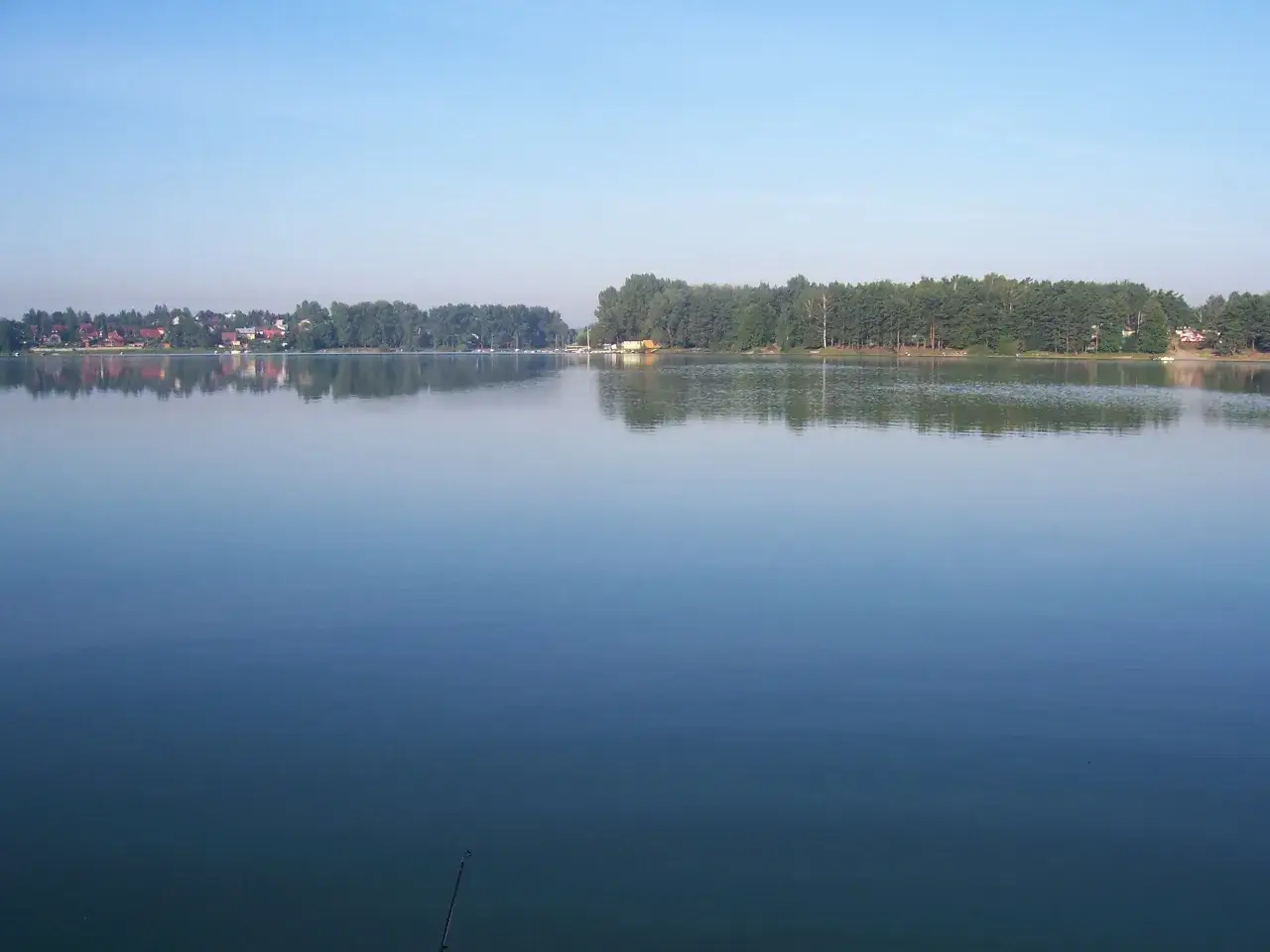 Spokojne jezioro Przeczyckie odbija błękitne niebo i zielony las. Na horyzoncie widać zabudowania i łodzie.