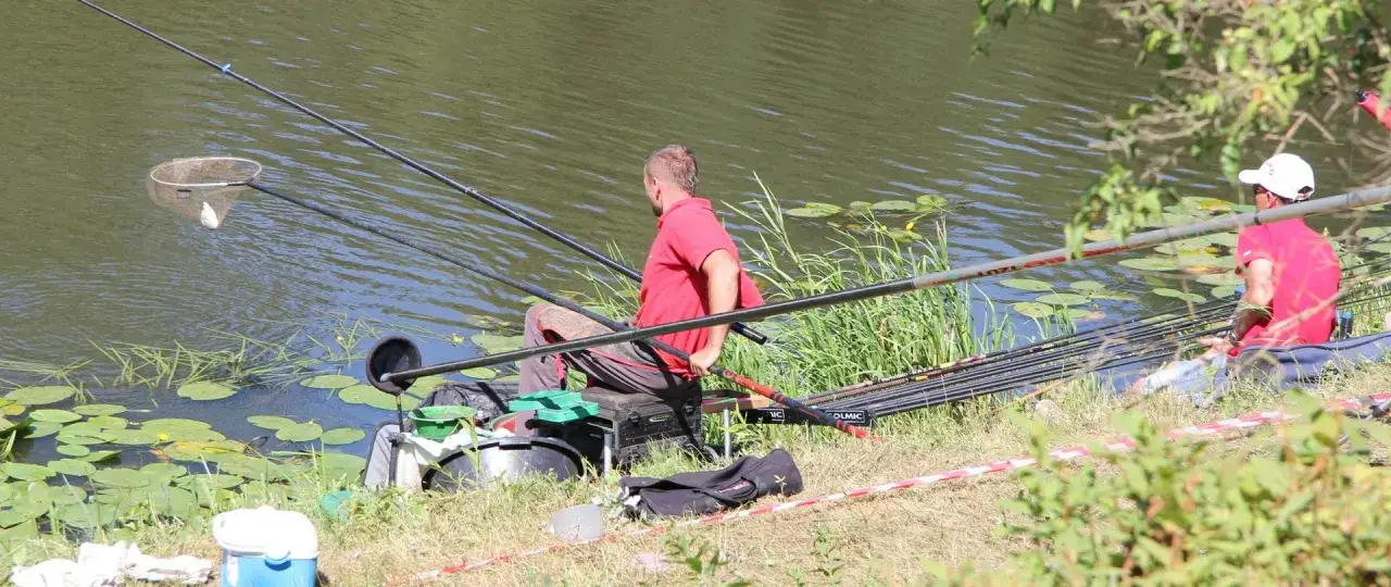 Dwóch wędkarzy siedzi nad wodą, jeden z nich właśnie wyciąga rybę z sieci. W tle widać spokojny zbiornik zaporowy z liliami wodnymi.